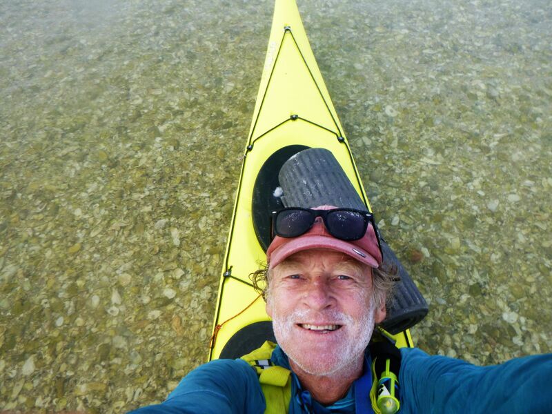 A selfie shows a man with sunglasses and a red cap, smiling in front of a yellow kayak. He is wearing a blue long-sleeved shirt and a yellow life vest. The kayak is on clear water, and the background shows the sandy bottom of the water. The man's face is the main focus, with the kayak and water providing context to his outdoor activity.